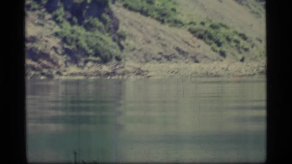Glacier Bay, Alaska home-movie frame showing calm water beneath a rocky, brushy slope with faded color and film marks.