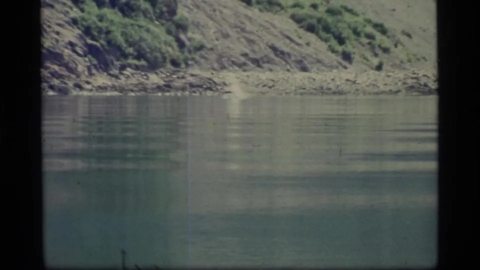 1977 8mm Alaska shoreline view with broad calm water, a steep rocky bank, green brush, and visible home-movie grain.