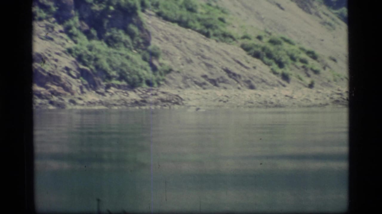 Calm lake water below a steep rocky hillside with green vegetation.