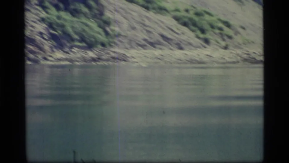 Glacier Bay lake surface and rocky shore appear in soft 1970s 8mm color, with a small distant dark shape near the water.