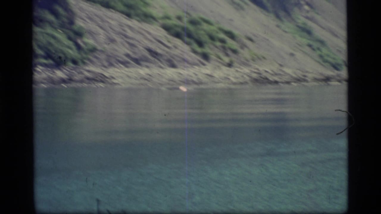 1977 Glacier Bay home-movie frame showing still water, rocky shoreline, and a steep green-gray mountainside.