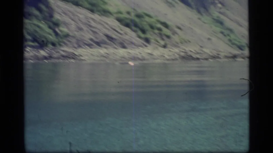 1977 Glacier Bay home-movie frame showing still water, rocky shoreline, and a steep green-gray mountainside.