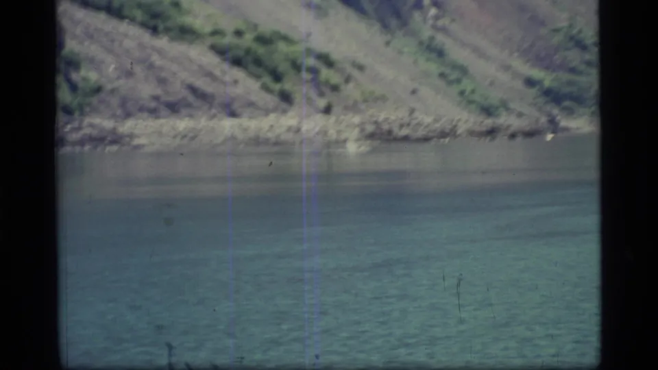 1977 Glacier Bay frame with reflective water, a rocky shore, and a tree-dotted slope under natural light.