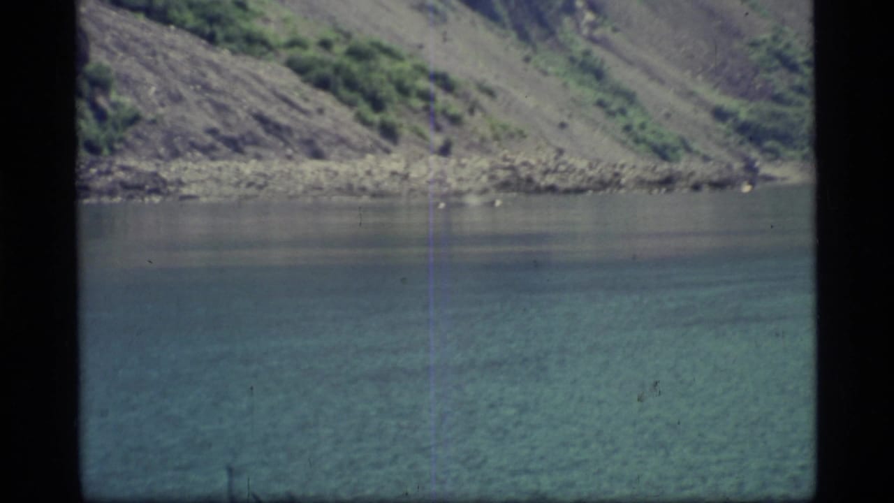 1977 8mm Glacier Bay view of calm blue-green water meeting a steep rocky mountainside with sparse green vegetation.