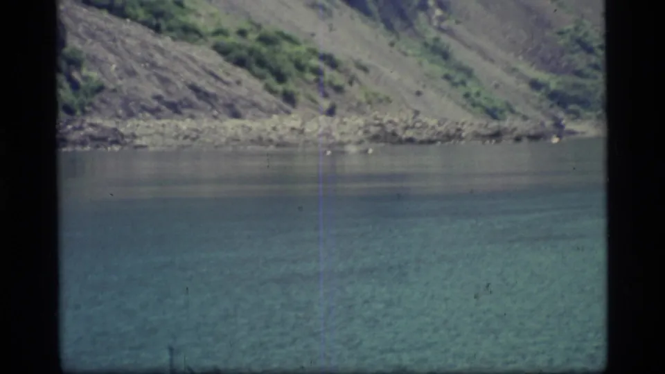 1977 8mm Glacier Bay view of calm blue-green water meeting a steep rocky mountainside with sparse green vegetation.