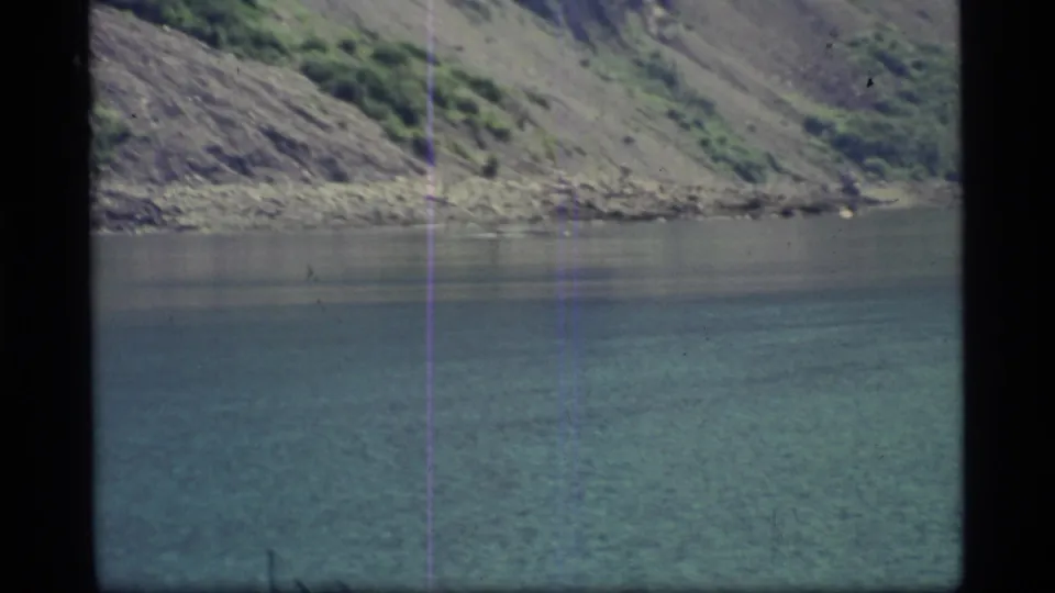 1977 8mm Alaska home-movie frame showing blue-green Glacier Bay water, a rocky shore, and a steep mountain slope.