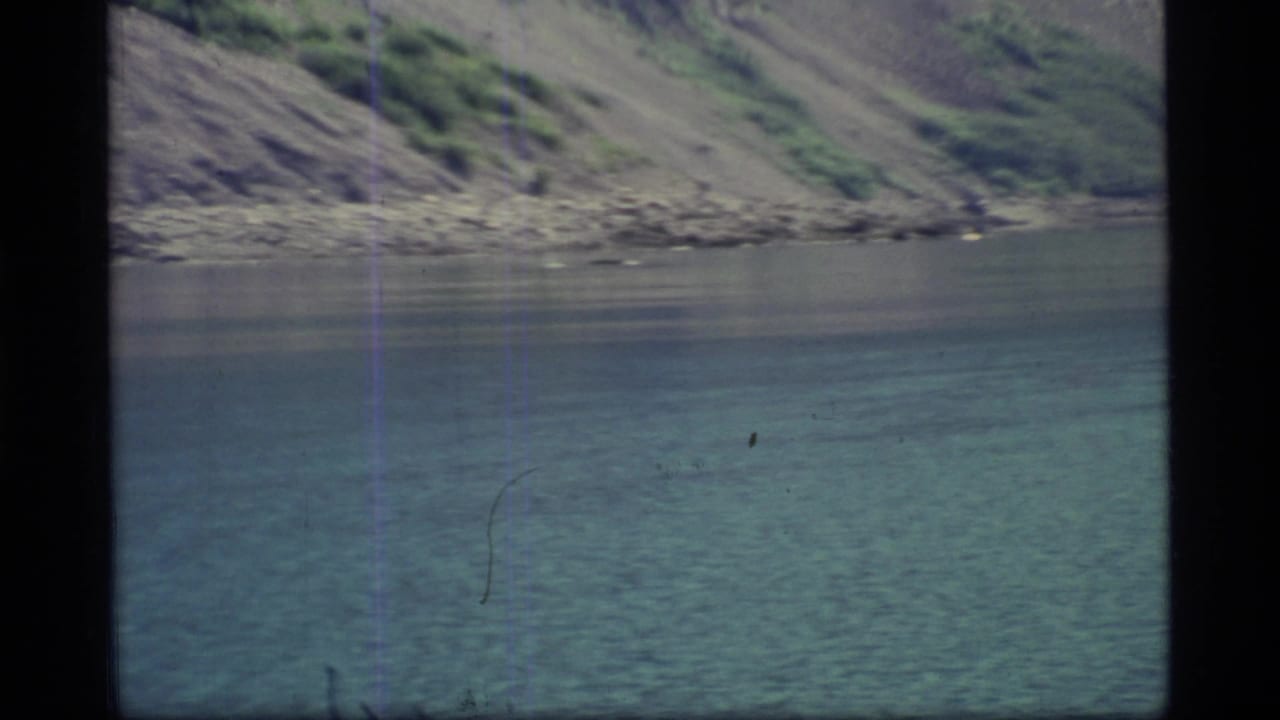 Calm turquoise water below a steep rocky shoreline with brush-covered slopes.