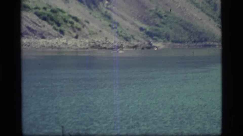 A quiet Glacier Bay shoreline is seen across still turquoise water, with soft 8mm grain and dark frame edges.
