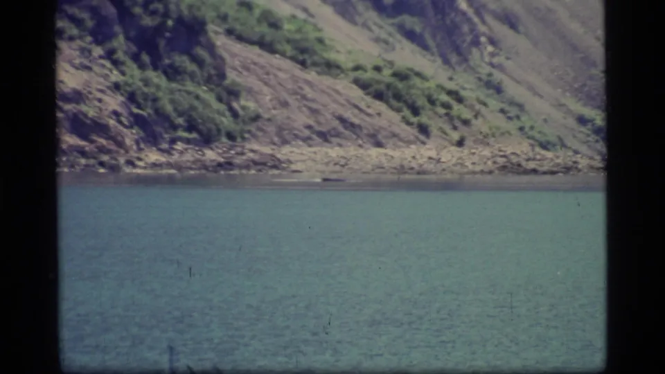 The 1977 home-movie frame holds on Glacier Bay water with a narrow rocky shore and brushy hillside in the background.
