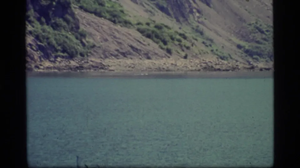 Glacier Bay, Alaska, appears in a quiet 1977 8mm frame with calm water, rocky shoreline, and sparse green vegetation on the slope.