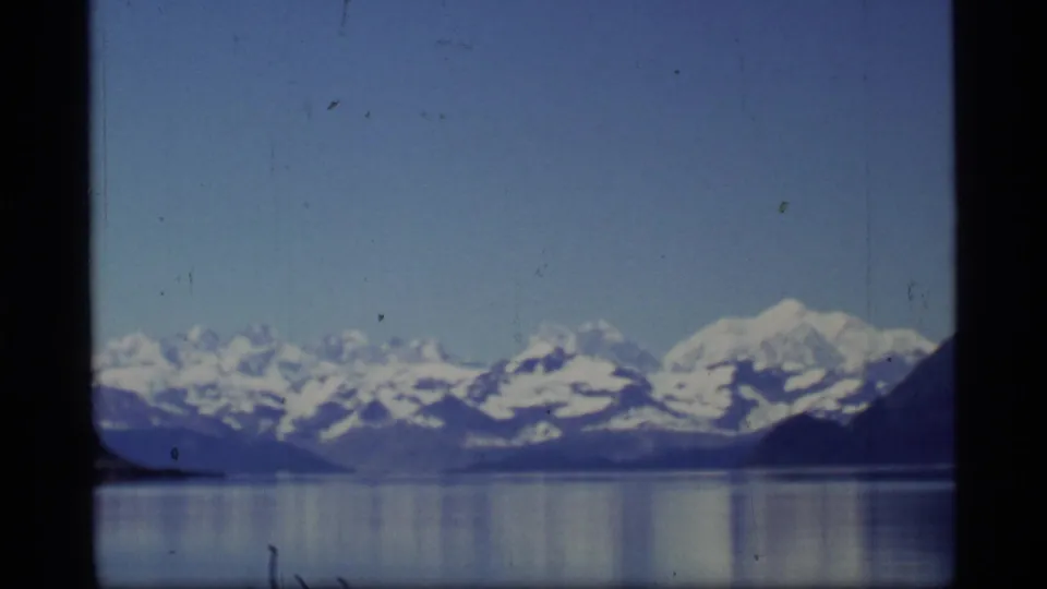 A wider 1977 Glacier Bay view shows snow-covered mountains reflected in calm water beneath a clear blue sky, with 8mm grain and edge darkening visible.