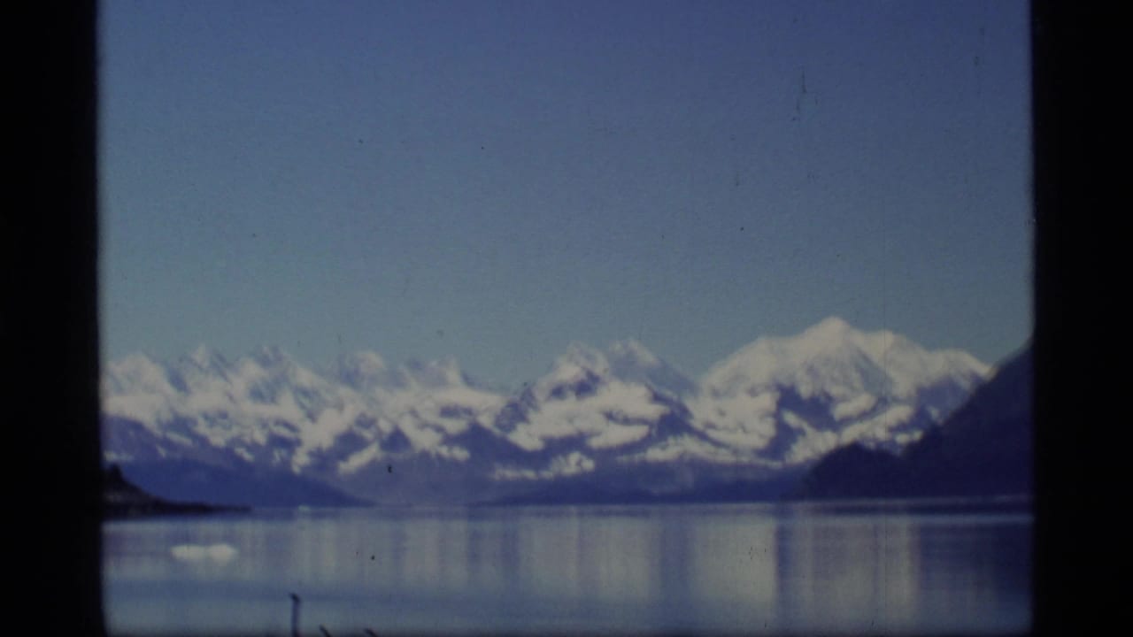 1977 Glacier Bay, Alaska 8mm frame showing distant snow-covered mountains and reflective water through a dark divided window view.