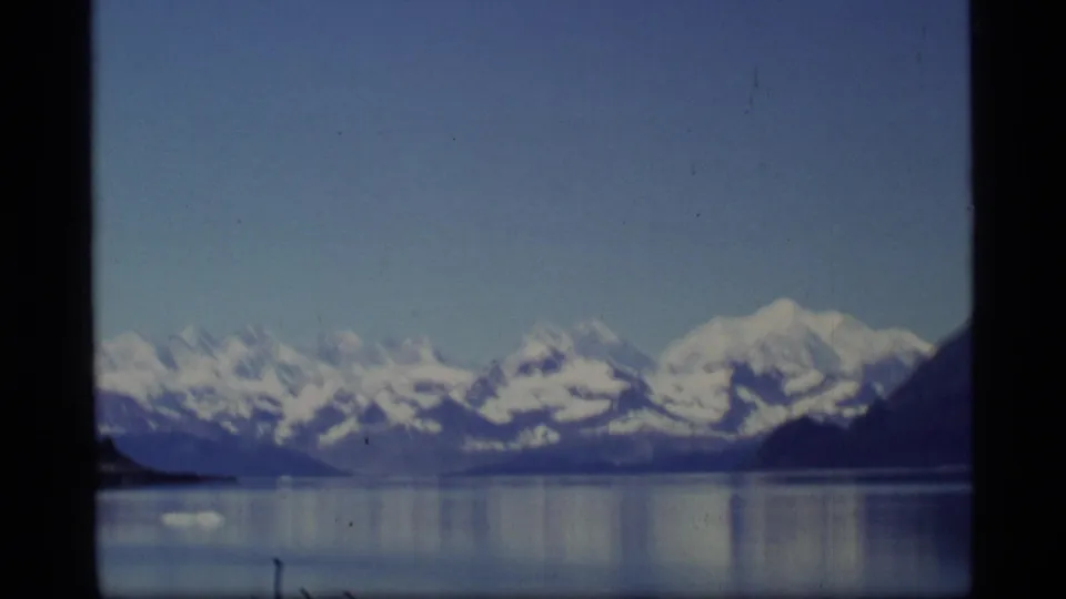 1977 Glacier Bay, Alaska 8mm frame showing distant snow-covered mountains and reflective water through a dark divided window view.