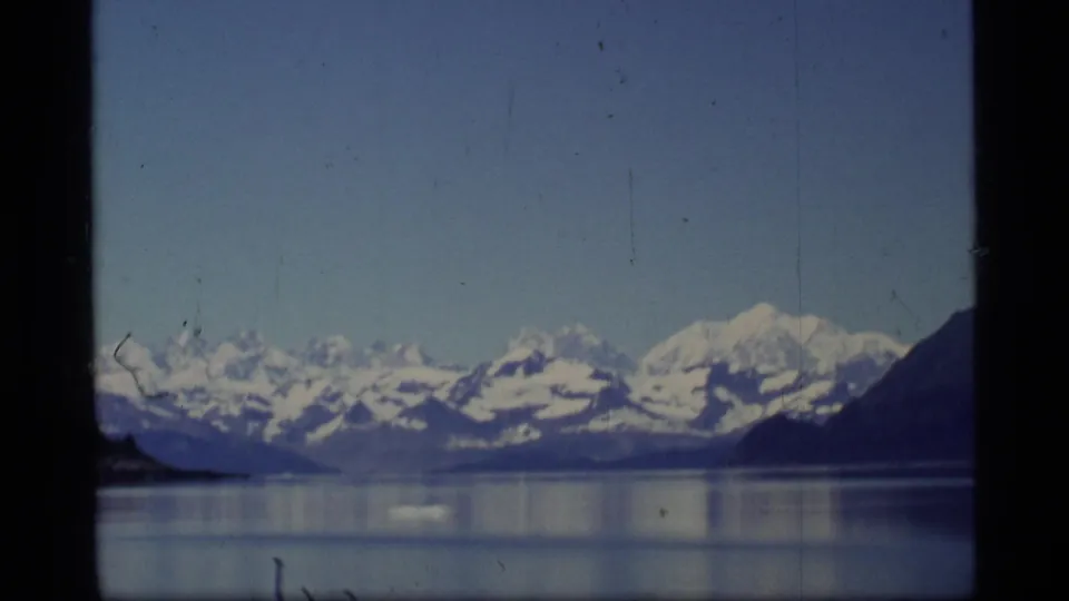 Glacier Bay, Alaska home-movie frame with snowy peaks, still water, pale sky, and dark vertical window framing.