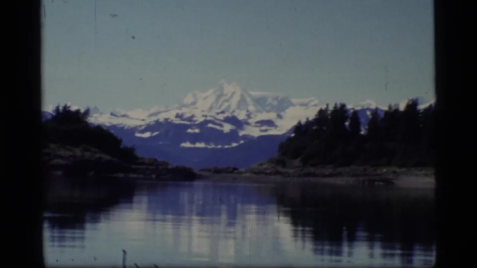 1977 Glacier Bay scenic frame showing a snow-covered mountain beyond dark shoreline trees and quiet reflective water.