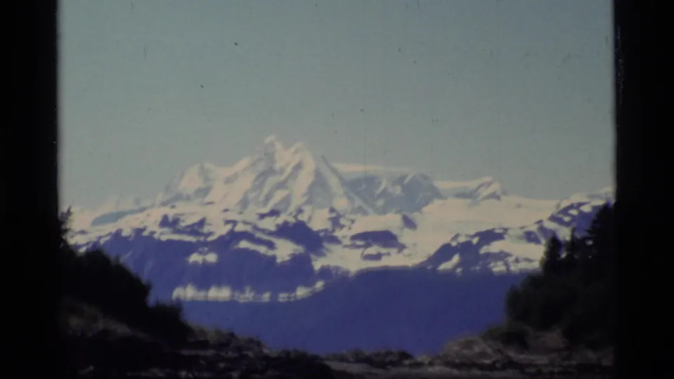 1977 8mm Glacier Bay view of a snow-capped mountain rising beyond a dark rocky shoreline under a pale sky.