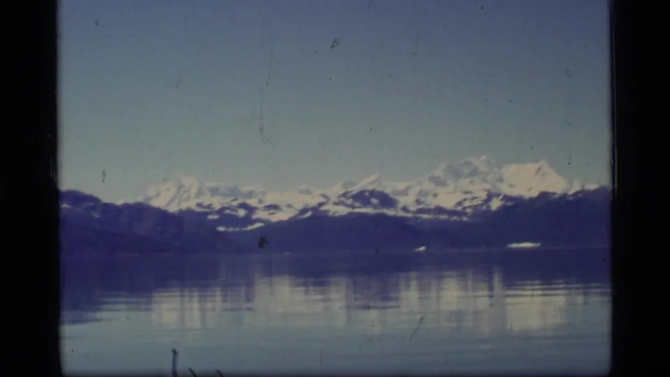 1977 8mm Glacier Bay landscape with distant snow-covered mountains mirrored in still bay water.