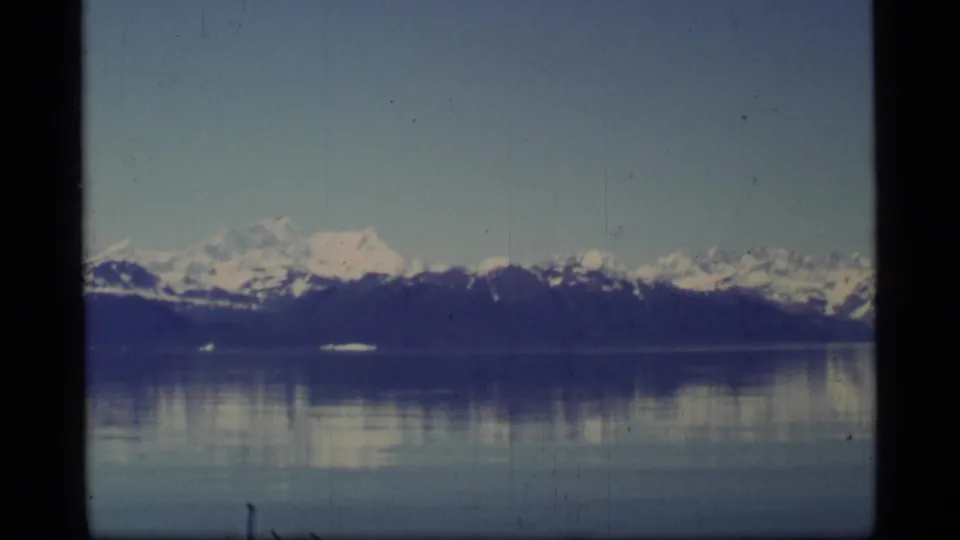 1977 8mm Glacier Bay view of snow-capped peaks across still water, with muted reflections and film grain.