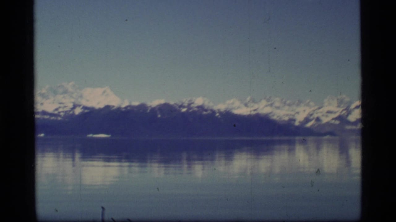 Distant snow-capped mountains reflected in calm water beneath a blue sky.