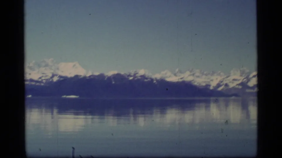 1977 Glacier Bay 8mm frame showing snow-capped mountains and dark shoreline reflected across still water.