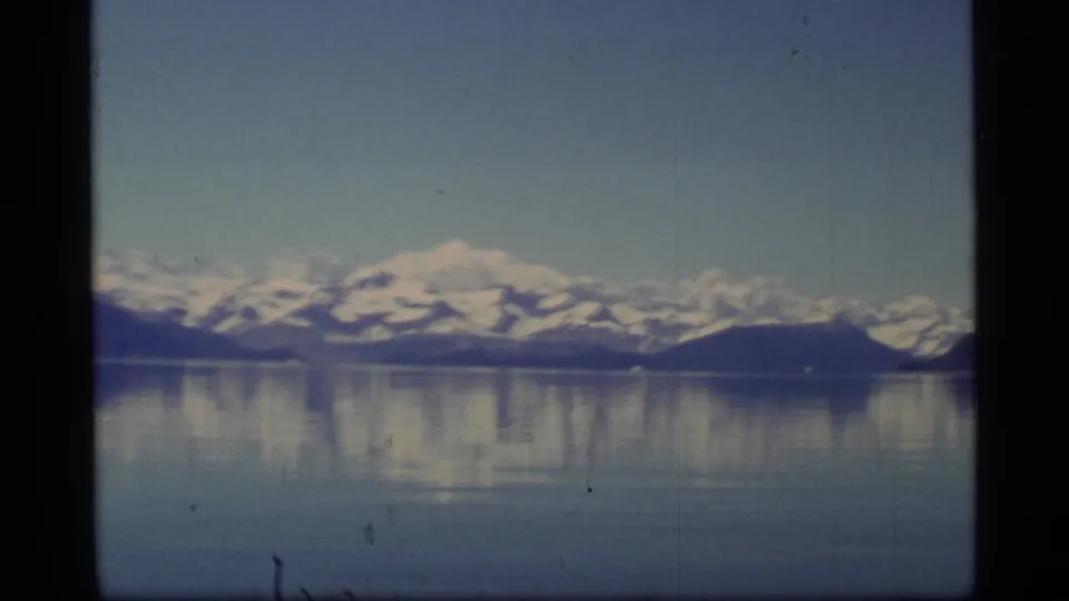 1977 Alaska home-movie frame showing Glacier Bay mountains, a low dark headland, and mirrored water.