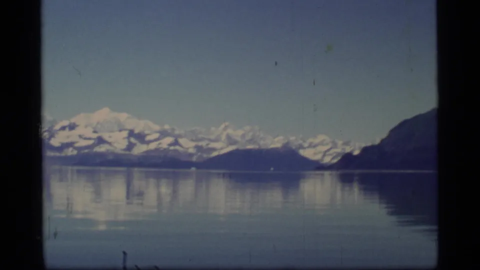 Glacier Bay 8mm frame with snowy mountains, island-like landforms, a dark right-hand slope, and still reflective water.