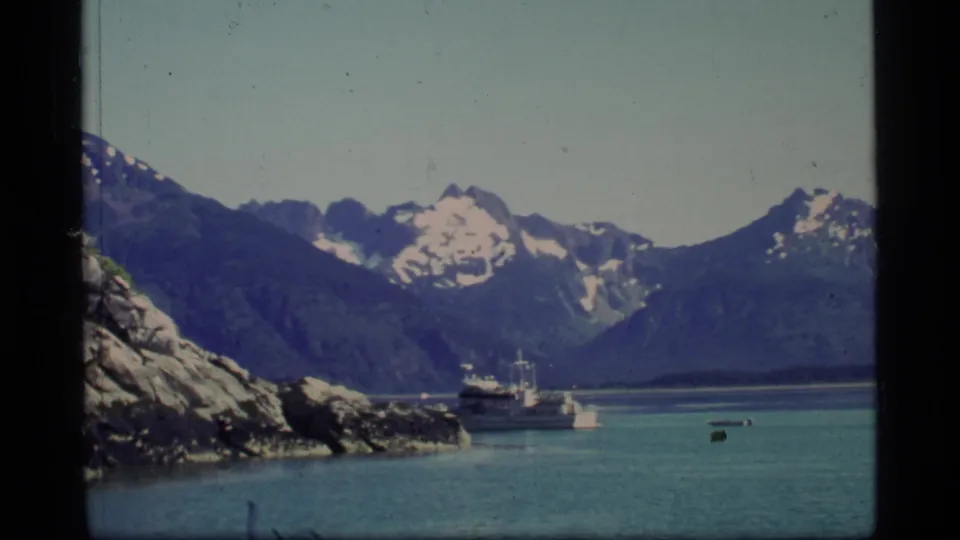 1977 Glacier Bay view of a ferry on blue-green water, with rocky shore and snow-capped mountains seen through a vessel window.