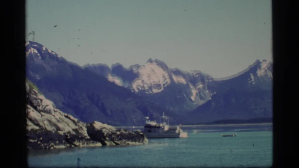 1977 Glacier Bay fjord scene with a ferry near rocky shoreline, blue-green water, and snow-capped mountain peaks in the distance.