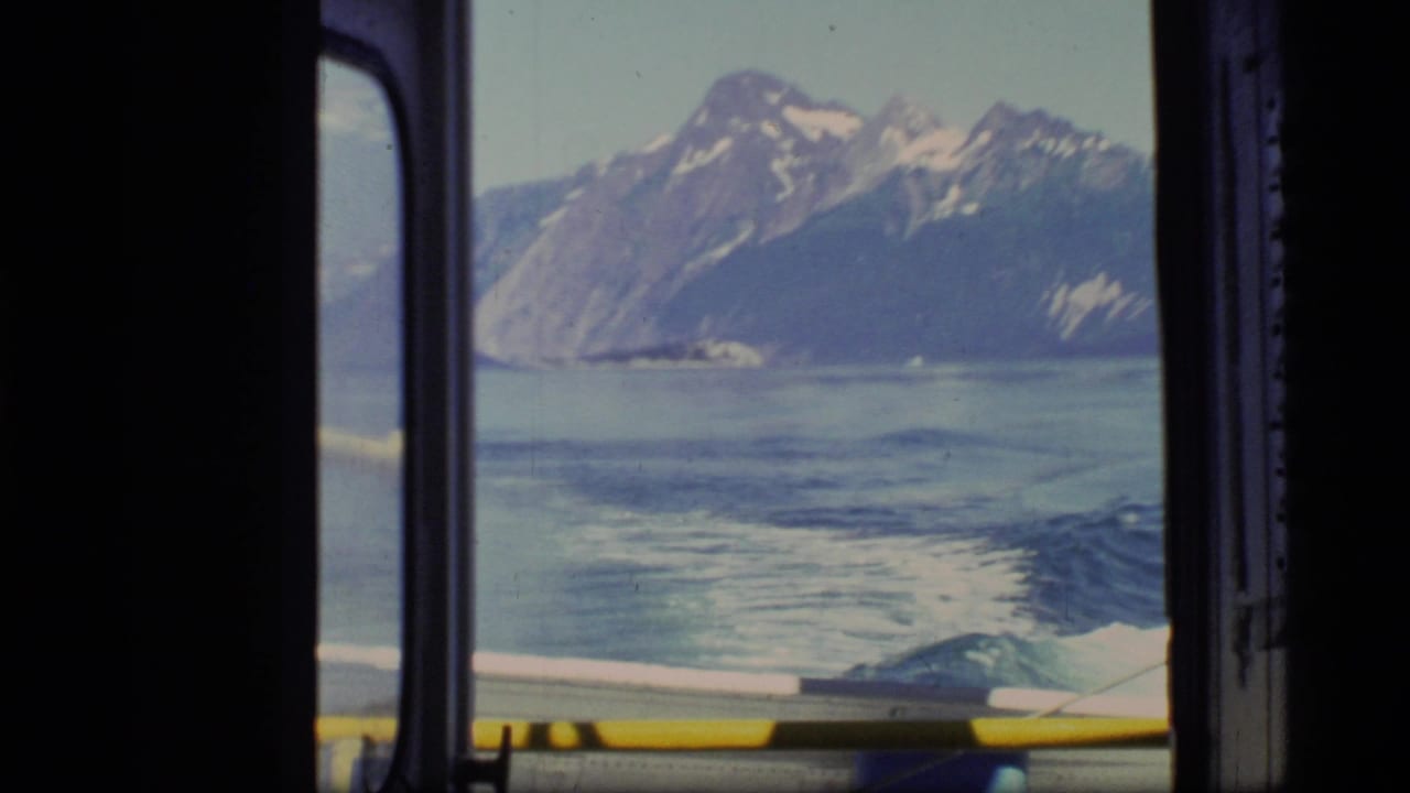 Mountain and bay view through a dark boat window with wake trailing below.