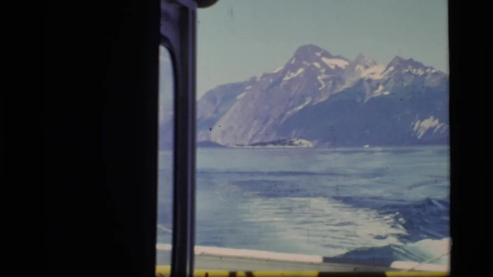 1977 Glacier Bay footage frames mountains across the bay from inside a moving boat, with wake visible near the lower edge.