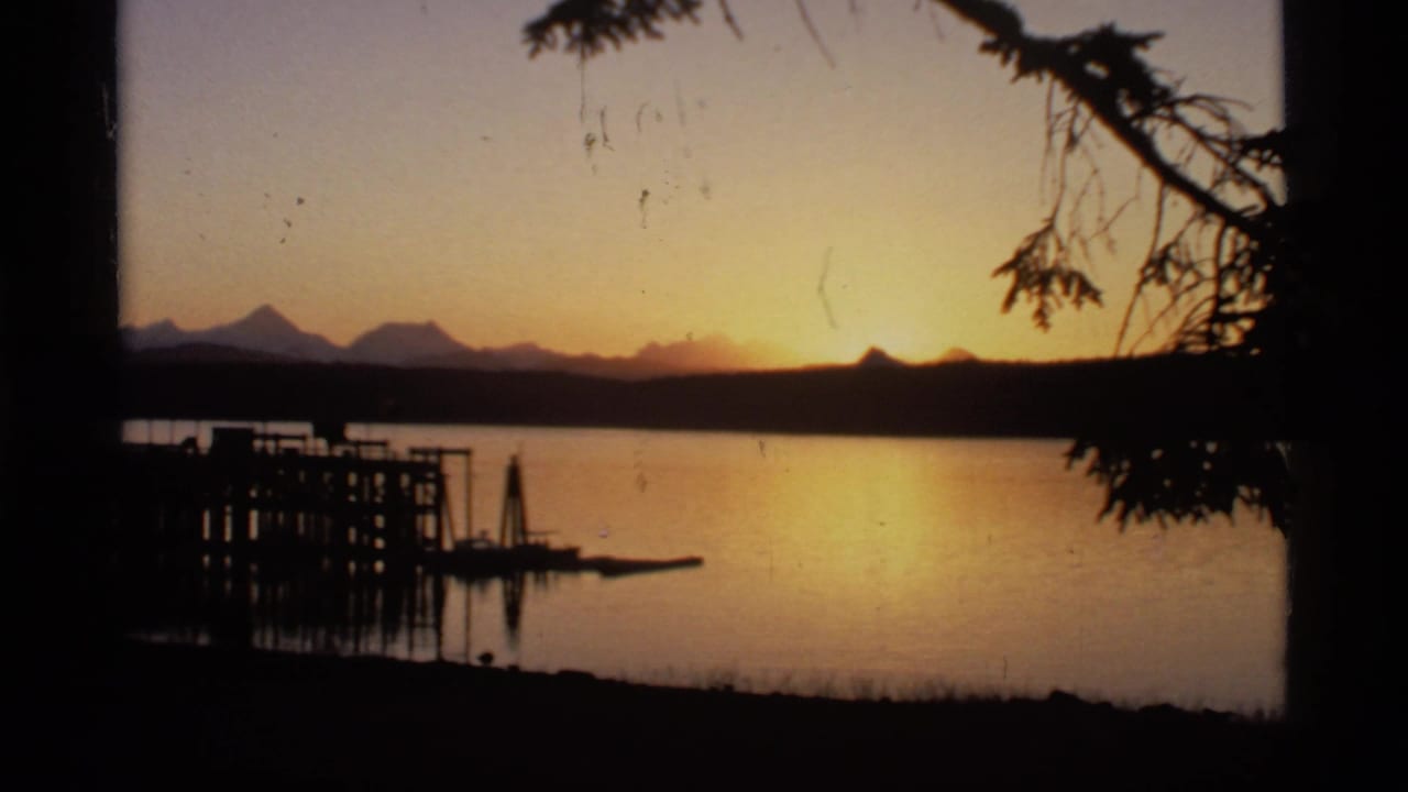 Sunset over a calm lake with distant mountains, a dock silhouette, and dark tree branches.