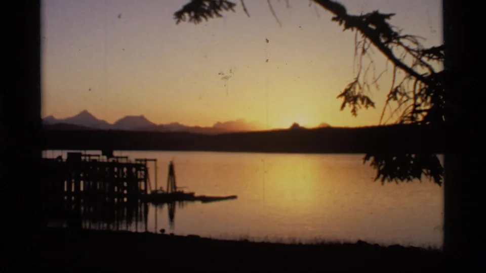 1977 Glacier Bay, Alaska 8mm frame with golden sunset reflected on lake water, distant mountains, dock structure, and dark tree limbs.