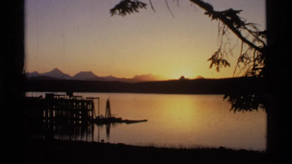 1977 Glacier Bay, Alaska 8mm frame showing a low sunset, still water, mountain silhouettes, dock forms, and foreground tree branches.