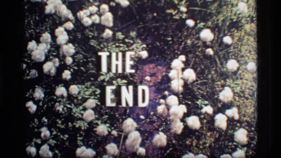 1977 8mm frame of a floral THE END title card with white letters, blossoms, and dark foliage.