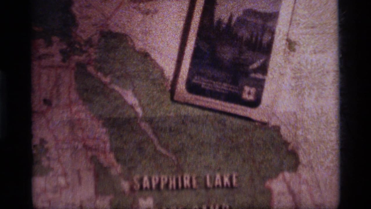 Faded 8mm frame from the Sapphire Lake clip showing a map surface, partial Sapphire Lake lettering, and a small photograph or card above it.
