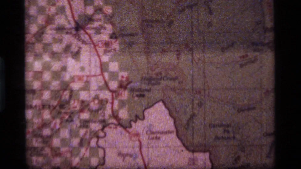 1970s 8mm map frame with a red route line crossing labeled places, softened by grain, scratches, and warm faded color.