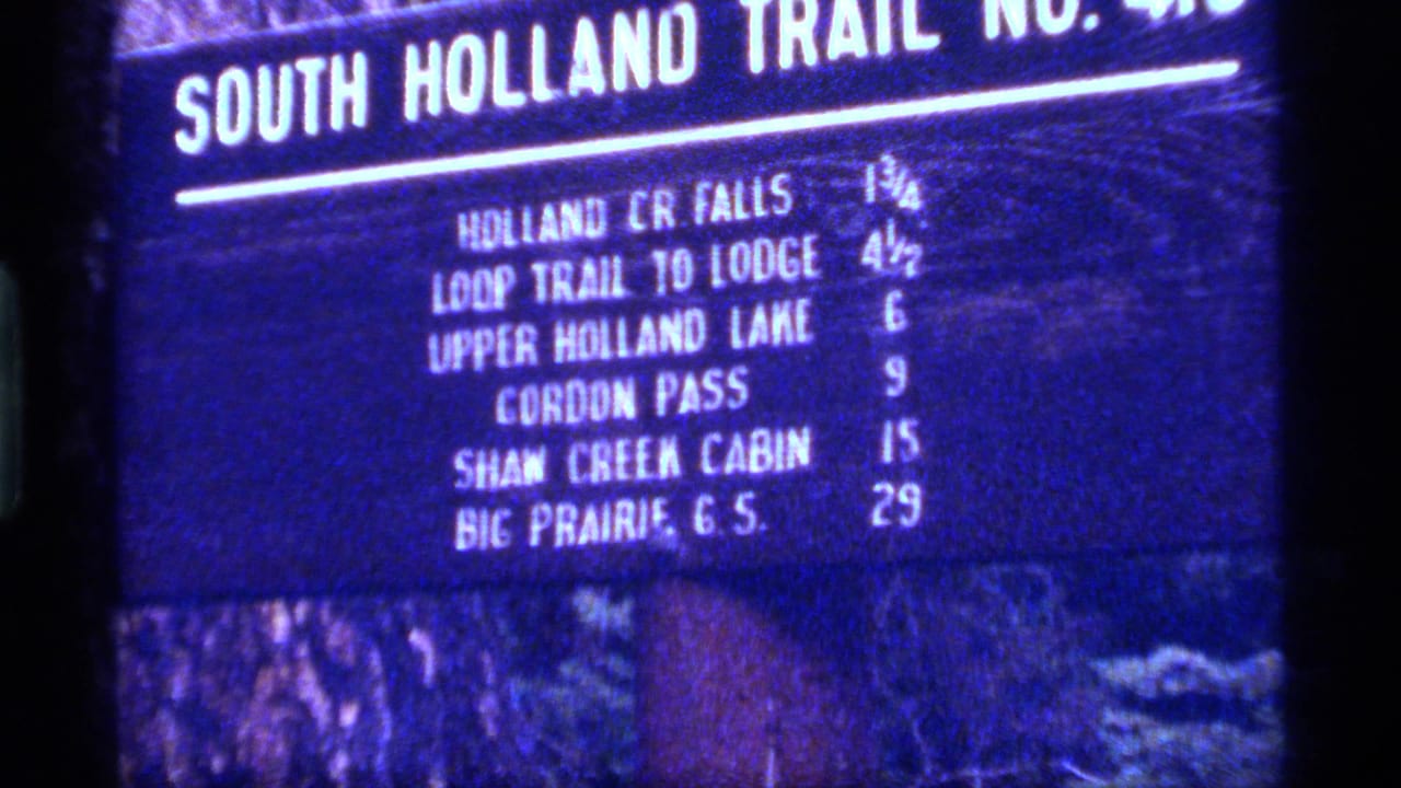 Blue-tinted 8mm frame showing a South Holland Trail No. 413 sign in a forested Montana setting, dated 1977 by metadata.