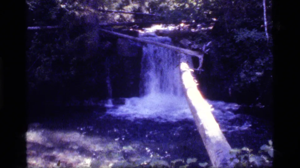 Archival 8mm frame showing the waterfall and angled log over a shaded Montana stream, with heavy blue color shift.