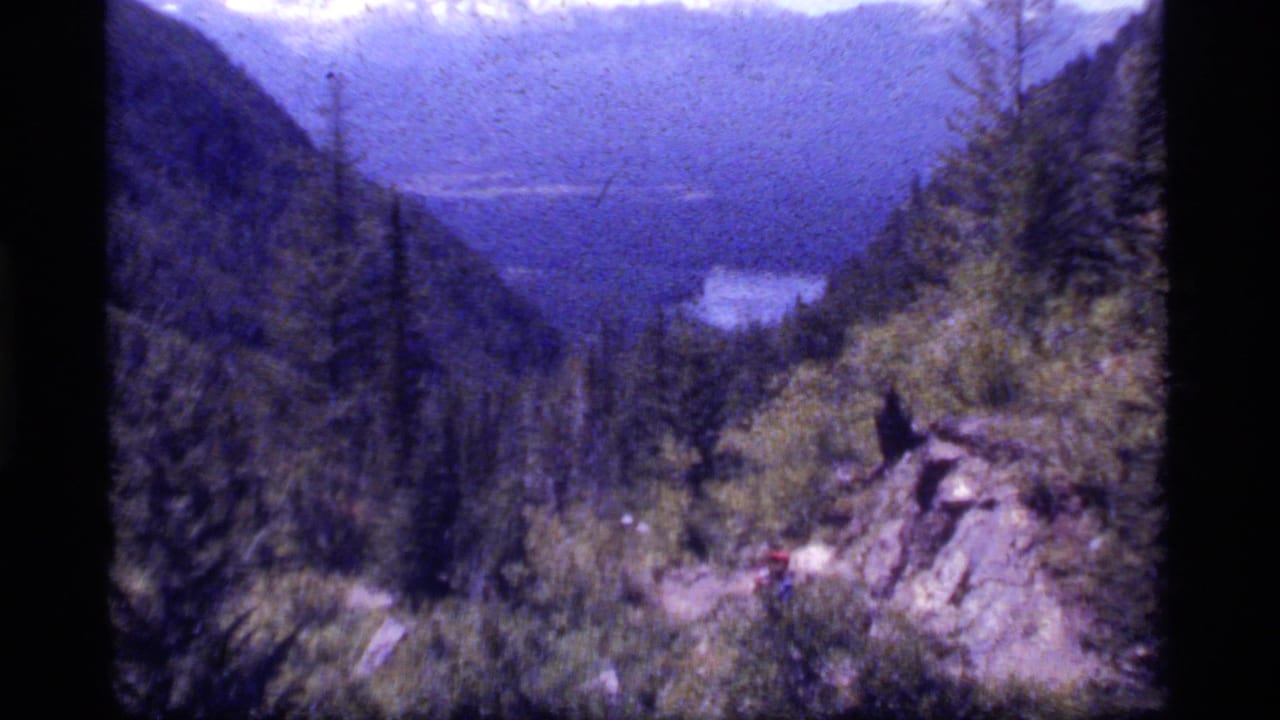 1977 8mm home-movie frame of a forested Sapphire Lake, Montana valley, with steep mountains, distant water, and a rocky overlook.