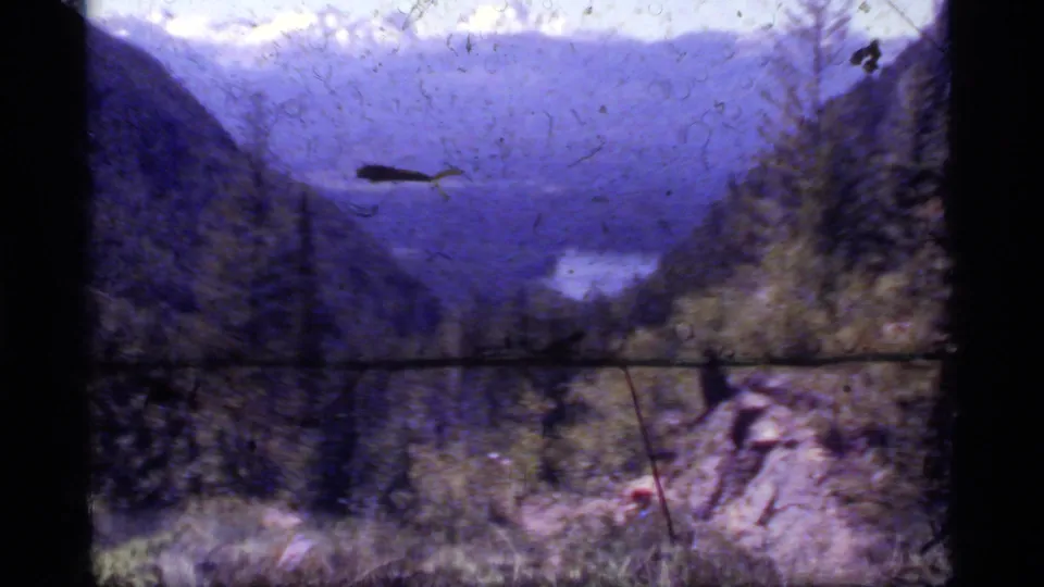 Sapphire Lake, Montana overlook on 8mm film, showing mountains, forest, distant water, and a small distant visitor-like figure near rocks.