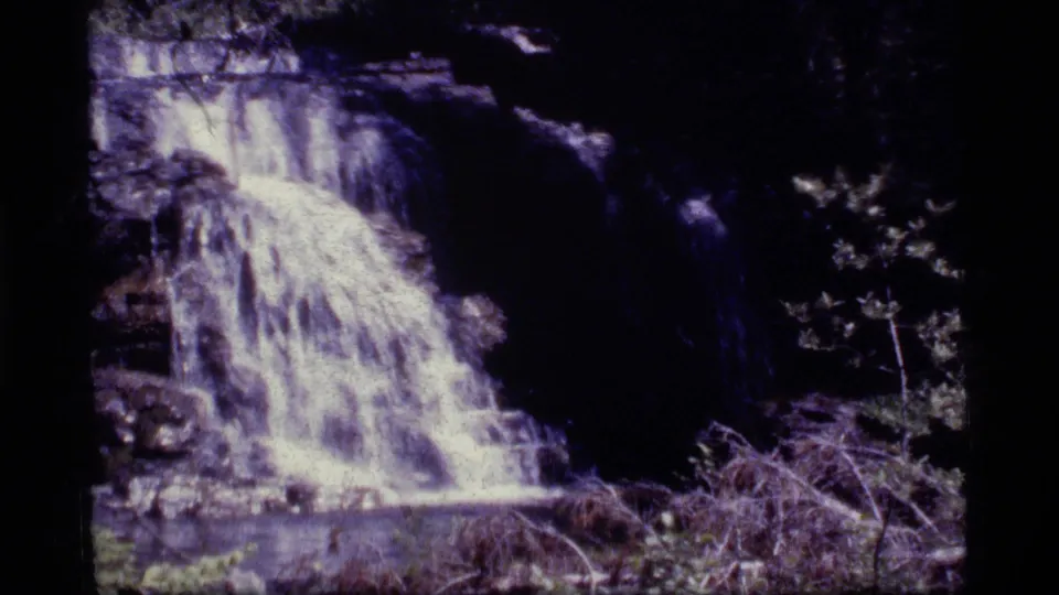 1977 8mm frame of a waterfall pouring over dark rock, with surrounding brush and forest shadow in Sapphire Lake, Montana footage.
