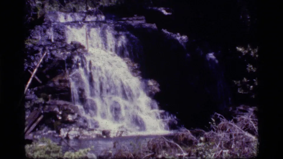 Vintage 8mm waterfall view from Sapphire Lake, Montana footage, with white water, dark rocks, brush, and dense shadowed trees.