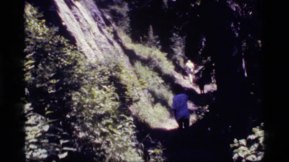 1977 Sapphire Lake, Montana 8mm frame showing a small figure in blue walking away on a sun-dappled forest path.