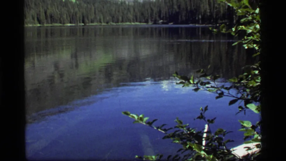 Sapphire Lake appears in a quiet 1977 8mm landscape view, with blue water, forest reflections, and shoreline branches.