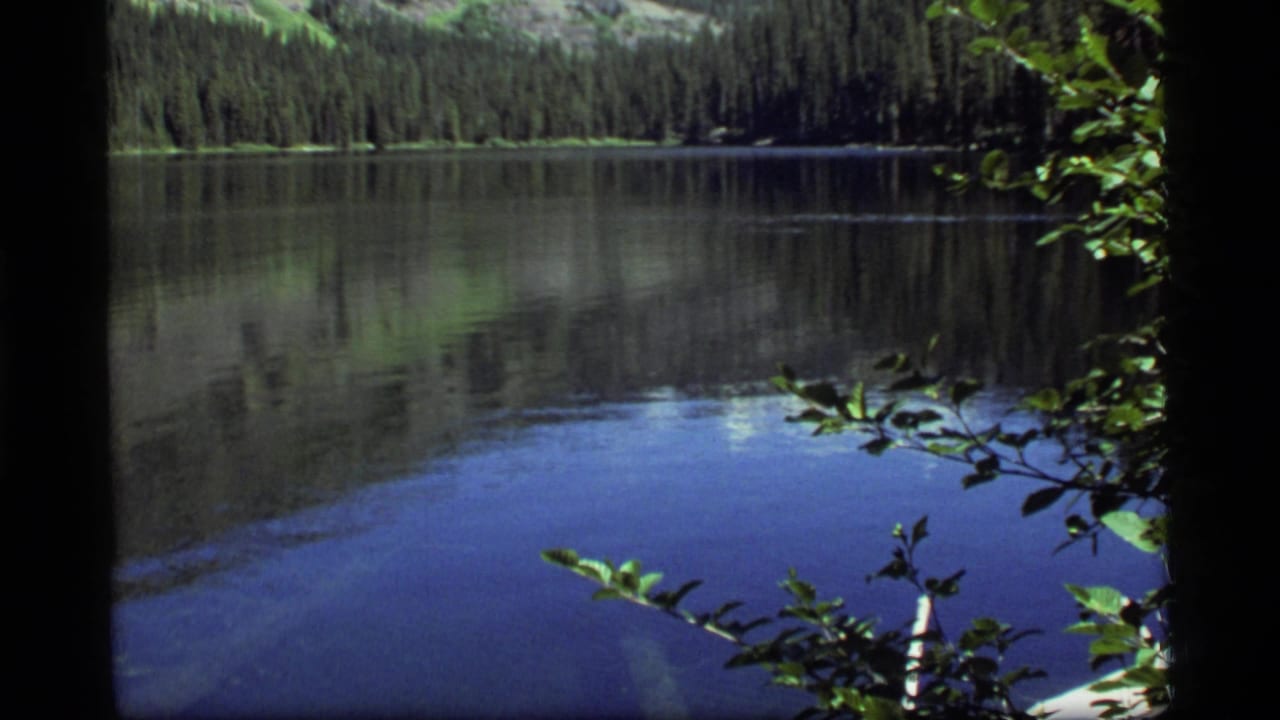1977 8mm view of Sapphire Lake in Montana, with still water reflecting evergreens and hillsides behind foreground leaves.