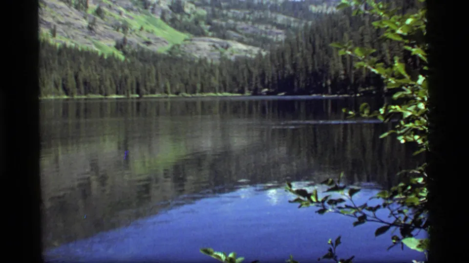 Vintage 8mm frame of Sapphire Lake, Montana, showing glassy water, conifer reflections, and leafy shoreline framing.
