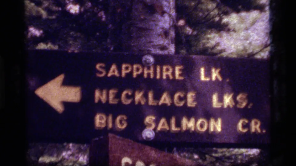 Readable Montana trail sign points left to Sapphire Lake, Necklace Lakes, and Big Salmon Creek in soft 1977 8mm footage.