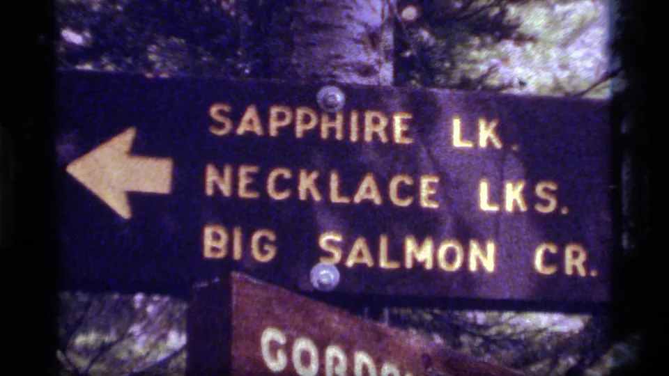 1977 Montana 8mm frame showing the Sapphire Lake trail sign from a handheld visitor viewpoint in a forested setting.