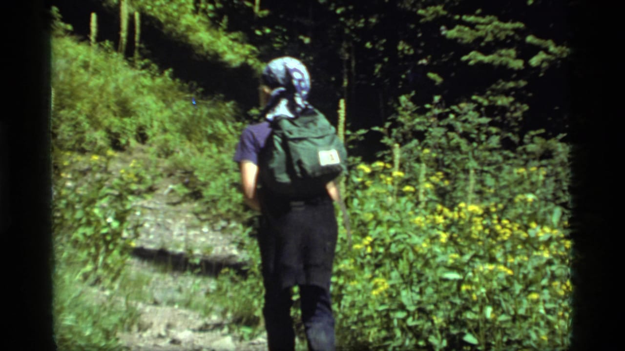 In 1977 Sapphire Lake, Montana footage, a lone hiker with a green backpack walks away along a shaded forest trail.
