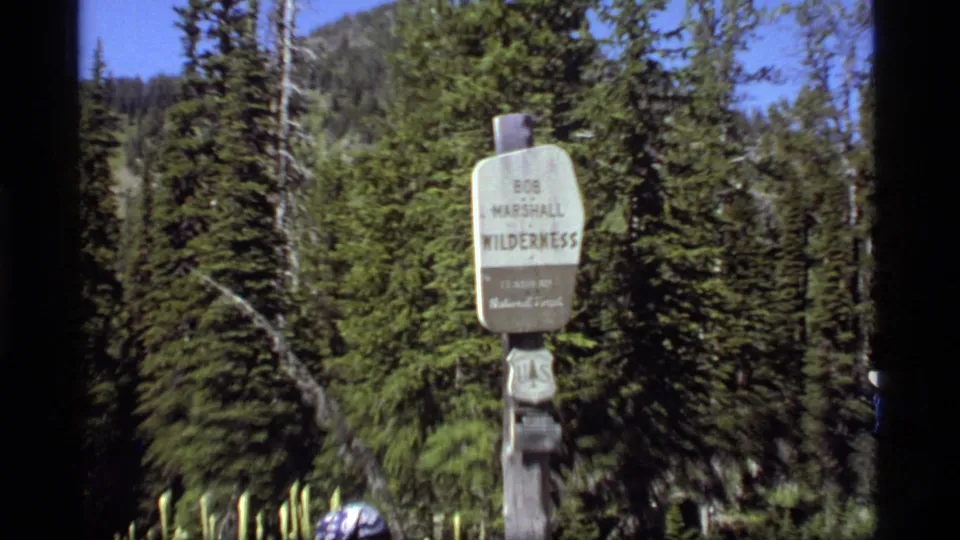1977 Montana home-movie frame with the Bob Marshall Wilderness sign and partial hikers at the margins.